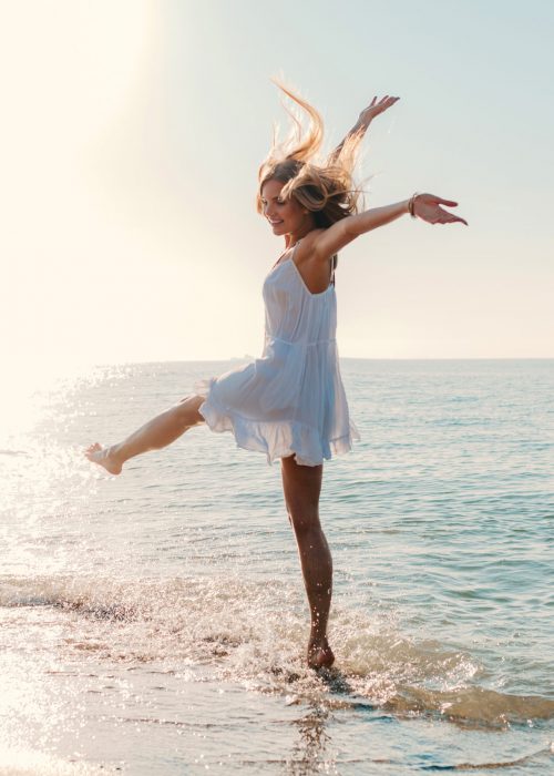 young attractive happy woman dancing turning around by sea beach sunny summer fashion style in white dress vacation
